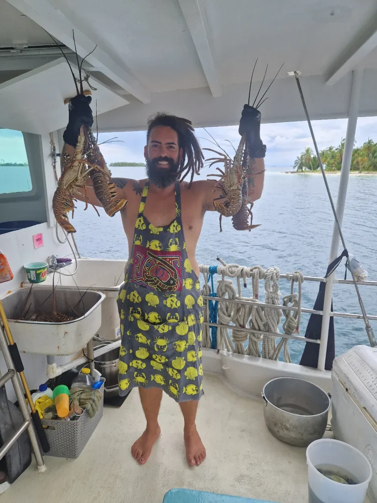 Captain Manu of Sovereign Grace holding up four fresh lobsters on deck, with a palm island behind him